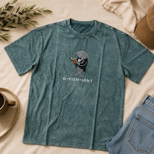 Può includere: Una t-shirt a maniche corte color verde acqua con lavaggio minerale, caratterizzata da una grafica di un pesce grigio che indossa uno smoking e un papillon neri mentre legge un libro aperto marrone. Sotto il pesce, il testo O-FISH-IANT.
