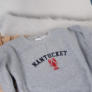 Puede incluir: Sudadera gris con la palabra "NANTUCKET" en azul marino y una langosta roja. La sudadera es de material suave. El dise&ntilde;o es de estilo cl&aacute;sico.