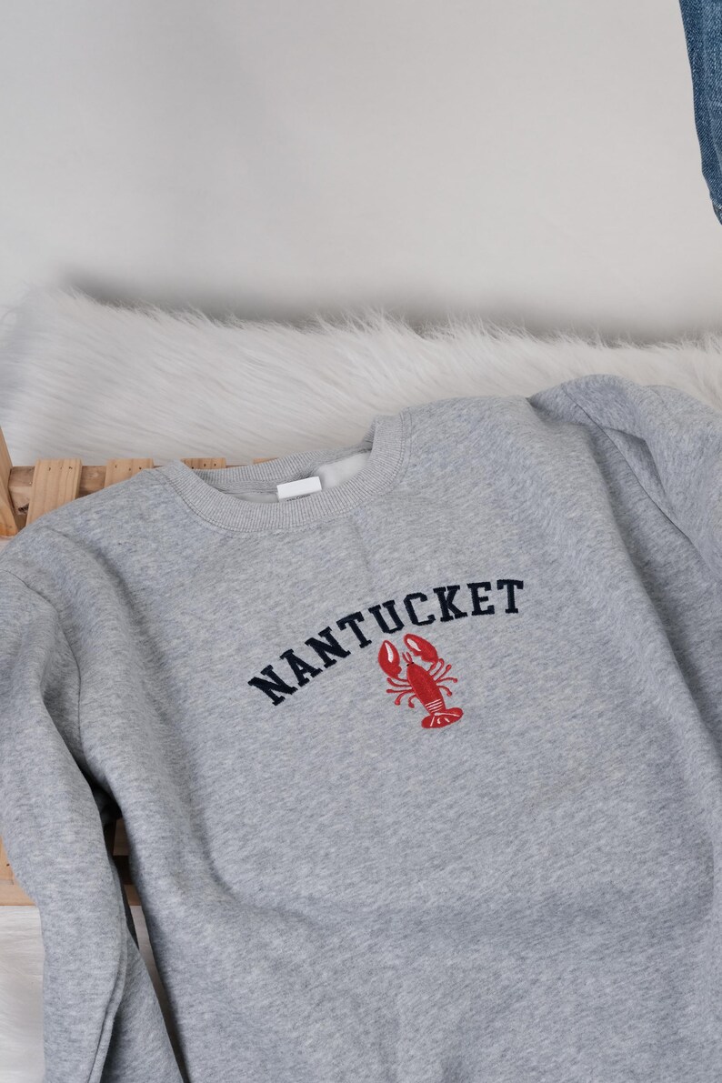Puede incluir: Sudadera gris con la palabra "NANTUCKET" en azul marino y una langosta roja. La sudadera es de material suave. El dise&ntilde;o es de estilo cl&aacute;sico.