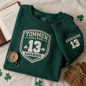 Puede incluir: Sudadera verde oscuro con texto bordado en blanco "Tommen College 13 Kavanaugh". La sudadera tiene cuello redondo y puños acanalados. También se ven una brújula, un libro y decoraciones de trébol.