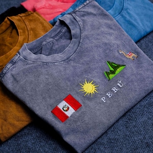 May include: Several stone-washed t-shirts in shades of grey, brown, pink, and blue. The grey t-shirt has embroidered designs of the Peruvian flag, a sun, mountains, a llama, and the word "PERÚ".