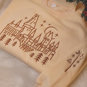 Peut inclure: Sweat-shirt crème avec une illustration marron d'un château, d'arbres, d'étoiles et d'un croissant de lune. Le motif est sur le devant et la manche du haut à col ras du cou et à manches longues. Le sweat-shirt est en matière douce.