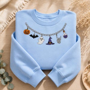 May include: A light blue crewneck sweatshirt featuring an embroidered silver chain graphic across the chest. The chain displays six Halloween-themed charms: an orange pumpkin, a black bat, a white ghost, a purple witch hat, a skeleton hand, and a potion.