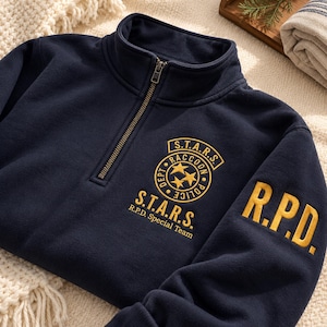 May include: A navy blue quarter-zip sweatshirt featuring yellow embroidered text and logos. The chest displays a circular S.T.A.R.S. Raccoon Police Dept emblem with three stars, while the sleeve features the bold yellow letters R.P.D.