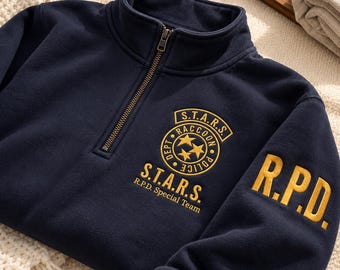 Può includere: Una felpa blu navy con mezza zip, caratterizzata da scritte e loghi ricamati in giallo. Sul petto è presente un emblema circolare S.T.A.R.S. Raccoon Police Dept con tre stelle, mentre sulla manica spiccano le lettere gialle R.P.D.