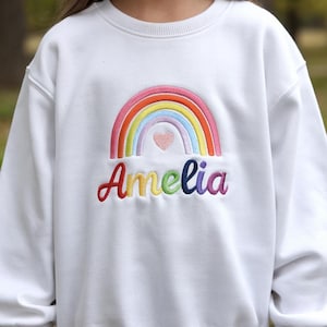 May include: White sweatshirt with a rainbow and a heart embroidered on the front. The name "Amelia" is embroidered below the rainbow in colourful letters.