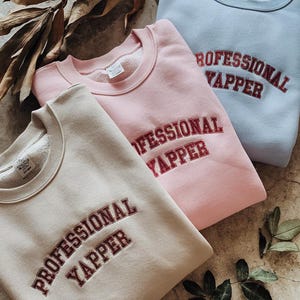 May include: Three sweatshirts in different colours: beige, pink, and light blue. Each sweatshirt has the text "PROFESSIONAL YAPPER" embroidered in maroon on the front.