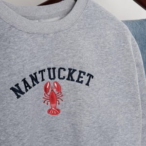 Puede incluir: Sudadera gris jaspeada con la palabra "NANTUCKET" bordada en azul marino sobre un gr&aacute;fico de langosta roja. La sudadera est&aacute; colgada de una percha de madera.