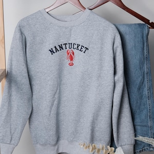 Puede incluir: Una sudadera gris con la palabra "NANTUCKET" bordada en azul marino sobre un gr&aacute;fico de langosta roja. La sudadera est&aacute; colgada en una percha de madera con unos vaqueros azules.