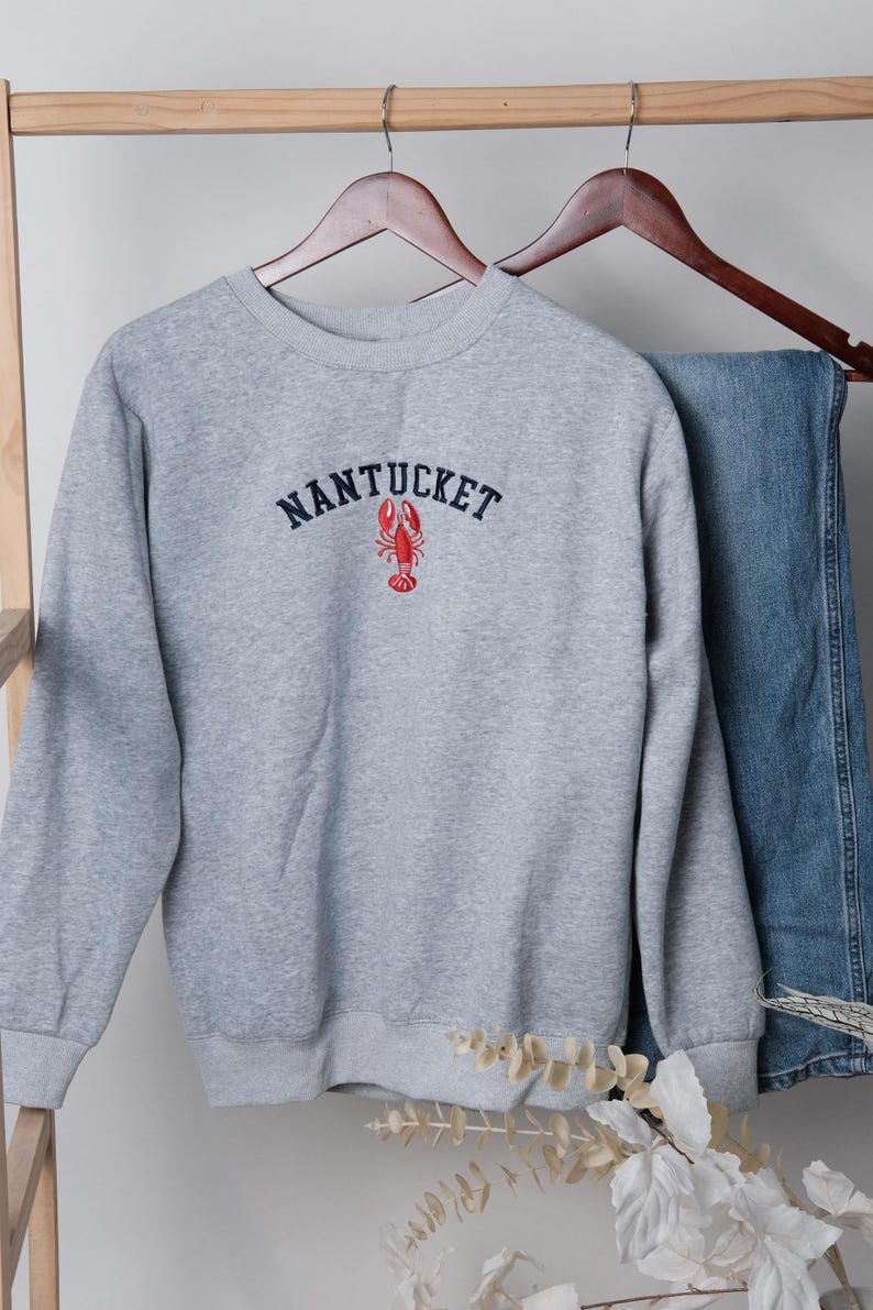 Puede incluir: Una sudadera gris con la palabra "NANTUCKET" bordada en azul marino sobre un gr&aacute;fico de langosta roja. La sudadera est&aacute; colgada en una percha de madera con unos vaqueros azules.