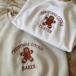 Może przedstawiać: Dwie bluzy z okrągłym dekoltem, jedna kremowa i jedna beżowa, z haftowanymi wzorami ludzików z piernika. Kremowa bluza ma napis "Christmas Cookie Tester", a beżowa "Christmas Cookie Baker". Haft jest w kolorze ciemnoczerwonym.