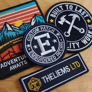 May include: Four embroidered patches: a square patch with a mountain sunset and the text ADVENTURE AWAITS, a circular patch with a large letter E and the text CUSTOM PATCH EMBROIDERED PATCHES, a circular patch with crossed hammers and BUILT TO LAST, and a rectangular patch with THELIEMS LTD.