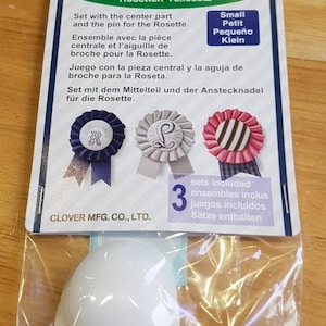 May include: A pack of three small rosette parts sets for crafts. The pack includes three rosettes, a centre piece, and a pin. The pack is labelled "Rosette Parts Set" and "Clover Mfg. Co., Ltd."