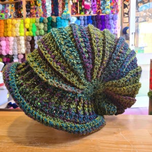 May include: A green, blue and purple crocheted hat with a spiral design. The hat is made with a variety of yarn colours and textures.