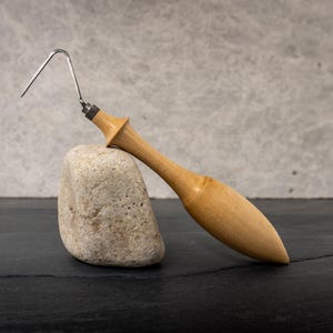 Puede incluir: Un instrumento dental de madera con un gancho de metal, apoyado sobre una pequeña piedra texturizada. El mango es de color marrón claro y el gancho es plateado. El fondo es gris.
