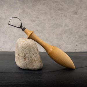 May include: A wooden tongue scraper with a stainless steel loop. The handle is light brown and has a smooth, tapered design. The scraper rests on a small, light-colored stone against a gray background.