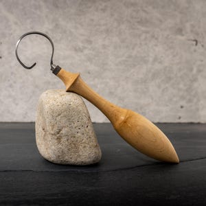 May include: A wooden carving tool with a curved metal blade, resting on a light-colored stone. The tool has a smooth, light brown wooden handle and a dark metal blade. The background is a textured gray surface.