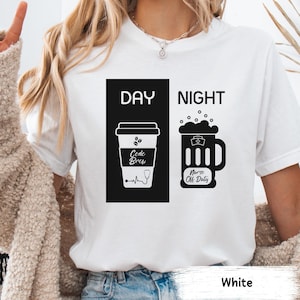 May include: White t-shirt with a black and white graphic design. The graphic shows a coffee cup with the text "Code Brew" and a beer mug with the text "Nurse Off Duty". The text "Day" is above the coffee cup and the text "Night" is above the beer mug.