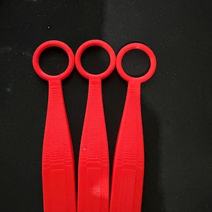 May include: Three bright red plastic tools, each with a ring handle and a textured, tapered blade. They are arranged in a row against a dark background. The design suggests a specialized function, possibly for crafting or repair.