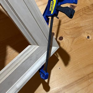 May include: A blue and black Irwin Quick-Grip bar clamp securing a light-coloured wooden frame with decorative moulding. The clamp features a yellow label with black text and a black metal bar, holding the mitered corner of the wooden frame together.