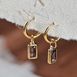 May include: A pair of gold hoop earrings with rectangular purple gemstone pendants. The earrings are displayed against a textured, off-white background, showcasing their polished finish and intricate design. The gemstones have a subtle, translucent quality.