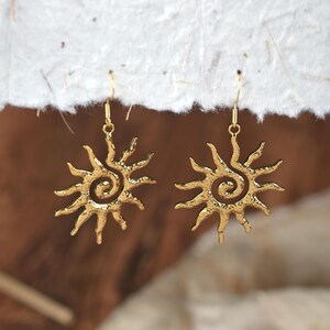 May include: A pair of gold-coloured sunburst earrings. Each earring features a detailed sun design with a spiral centre and radiating rays. The earrings are suspended from delicate gold-coloured hooks, showcasing a bohemian or celestial theme.
