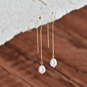 May include: A pair of gold-colored threader earrings with teardrop-shaped white pearls. The earrings hang from a delicate chain, with a small gold bar above the pearl. The background is a mix of a textured white surface and a wooden surface.