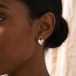 May include: Gold-colored heart-shaped stud earrings. The earrings have a padlock design with the word "FOREVER" engraved on the heart. The earrings are worn on a person's ear.