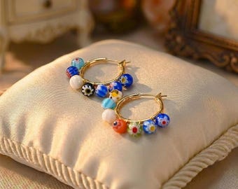 May include: A pair of gold hoop earrings adorned with colorful glass beads. The beads are in various colors, including blue, white, orange, and green, and are arranged around the hoops. The earrings are displayed on a cream-colored pillow.
