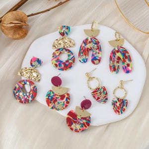 Pode incluir: Conjunto de brincos coloridos, abstratos e geométricos com detalhes dourados. Os brincos apresentam uma variedade de formas, incluindo círculos, arcos e semicírculos. Os brincos são feitos de argila polimérica e têm um efeito marmorizado.