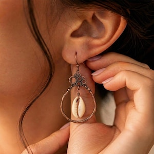 May include: A dangle earring featuring a hammered copper-toned metal hoop with a decorative circular cluster at the top. A single white cowrie shell hangs from the center of the hoop, suspended by a small wire-wrapped loop attached to the metal frame.