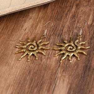 May include: A pair of gold-coloured sun-shaped earrings. Each earring features a spiral centre with radiating rays. The earrings have a hook closure and are displayed on a wooden surface.