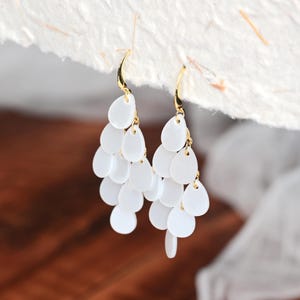 May include: A pair of white drop earrings with a cascading design. Each earring has multiple teardrop-shaped, white elements suspended from a gold-coloured hook. The earrings are designed to dangle and catch the light.