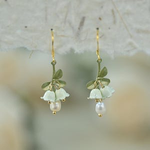 May include: Dangle earrings with a floral design. Each earring has a gold-coloured hook, a green stem with leaves, and three pale green bell-shaped flowers. A white pearl hangs from each earring.