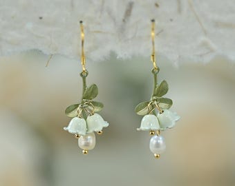 May include: A pair of dangle earrings featuring a floral design. Each earring has a gold-colored hook, a green stem with leaves, and three pale green bell-shaped flowers. A white pearl hangs from each earring.