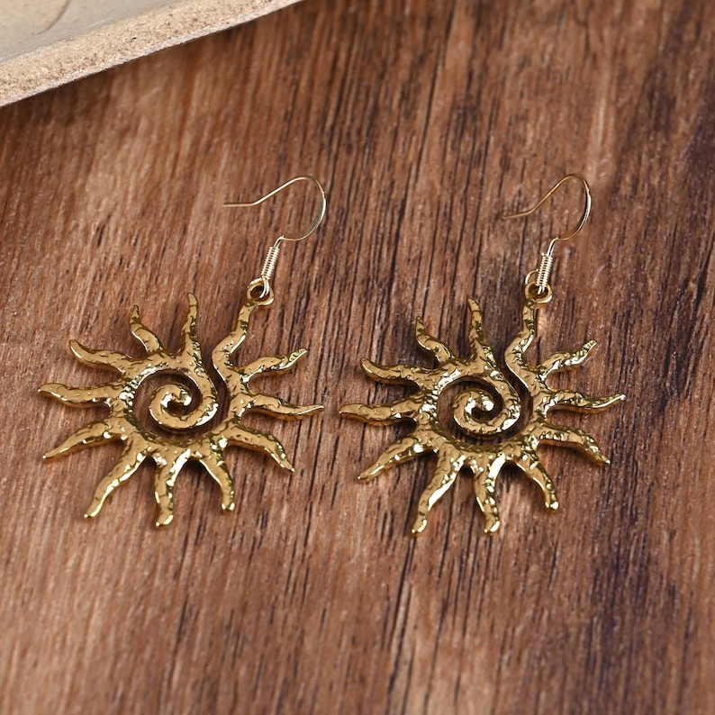 May include: A pair of gold-coloured sunburst earrings. Each earring features a sun design with a spiral centre and radiating rays. The earrings have a hook closure and are displayed on a wooden surface.