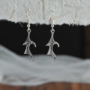 May include: A pair of silver-toned drop earrings featuring a detailed bat wing design. Each earring hangs from a standard silver-toned hook wire, showcasing textured, pointed wing structures with defined skeletal lines and a metallic finish.
