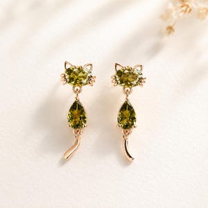 May include: A pair of rose gold-toned earrings shaped like cats. Each earring features an oval-cut olive green gemstone for the head, a pear-shaped olive green gemstone for the body, and a curved rose gold-toned metal tail dangling below.