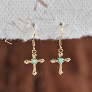 May include: A pair of gold-colored cross earrings with a turquoise-colored stone in the center of each cross. The earrings have a decorative design and are suspended from a simple hook.