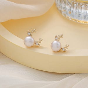 May include: A pair of gold earrings with a pearl and a small branch design. The earrings are on a white surface.