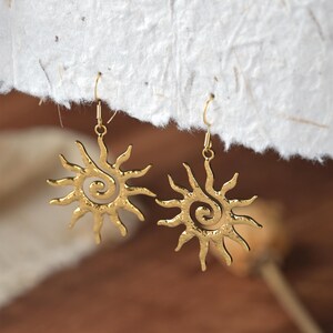 May include: A pair of gold-coloured sun-shaped earrings. Each earring features a spiral centre and radiating rays. The earrings are suspended from gold-coloured ear wires. The background is a soft, neutral colour.