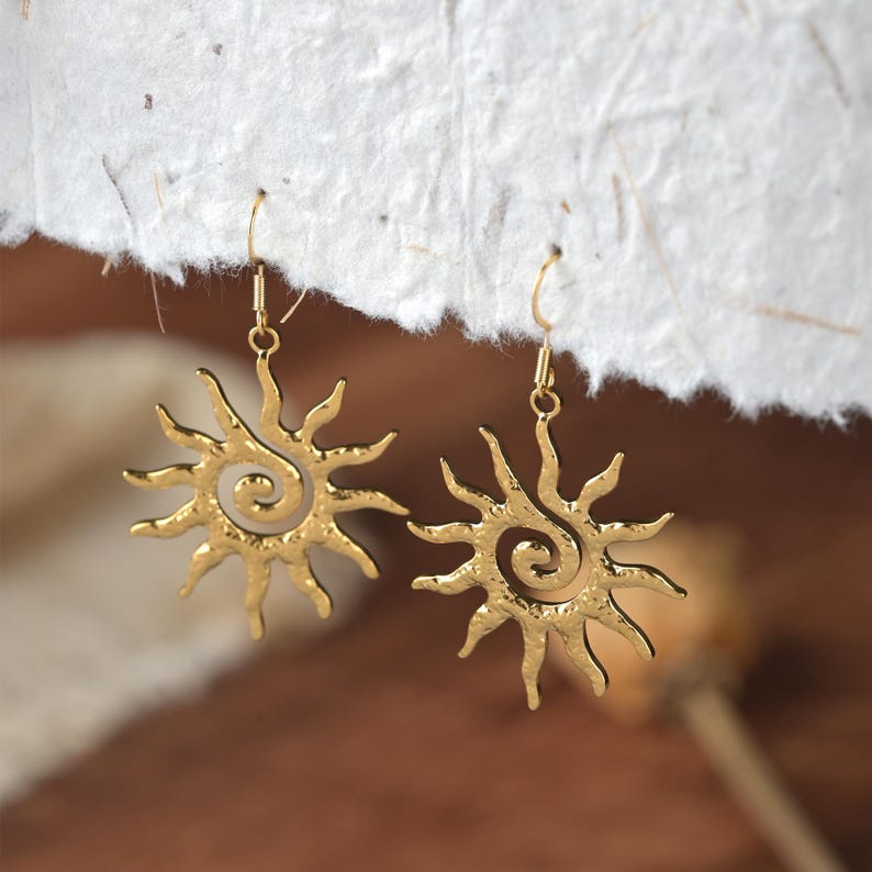 May include: A pair of gold-coloured sun-shaped earrings. Each earring features a spiral centre and radiating rays. The earrings are suspended from gold-coloured ear wires. The background is a soft, neutral colour.
