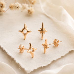 May include: Four gold-toned stud earrings featuring star-shaped designs. Two earrings are shaped like four-pointed stars with a circular cutout in the center, and two earrings are shaped like slender, elongated four-pointed stars with a cross-like center.