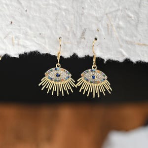 May include: A pair of gold-colored earrings with an eye design. The earrings feature a blue center surrounded by colorful gems and gold rays. The earrings are hanging against a textured white background.