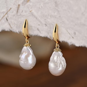 Può includere: Un paio di orecchini color oro con un gancio in metallo lucido curvo nella parte superiore e una perla bianca irregolare a forma di goccia che pende. La perla è fissata al gancio tramite una piccola calotta metallica decorativa smerlata.