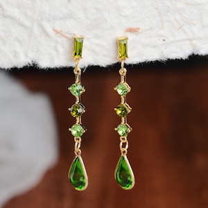 May include: Gold-toned dangle earrings with green gemstones. Each earring features a rectangular green stone at the top, followed by a series of round and square green stones, and a teardrop-shaped green stone at the bottom. The earrings are designed for pierced ears.