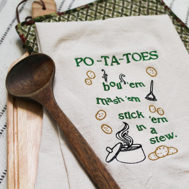 An unbleached off white linen tea towel embroidered with a quote from Lord of the Rings movie, the Two Towers. The Quote is Potatoes. Boil 'em, mash 'em, stick'em in a stew. Playful potatoes fall down to a  large whimsical pot, and a classic potatoes masher. The fonts represent the verbs, with steam coming off of the boil em phrase and the letters in mashed closely pushed together. Colors are a bold emerald green and a soft golden brown. Hang this towel in kitchen of any hobbit, elf, or wizard.