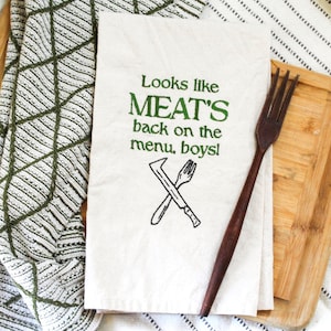 May include: Cream-coloured kitchen towel with green text saying "Looks like MEAT'S back on the menu, boys!" and a crossed knife and fork design. A dark wooden fork rests on the towel, next to a wooden chopping board.