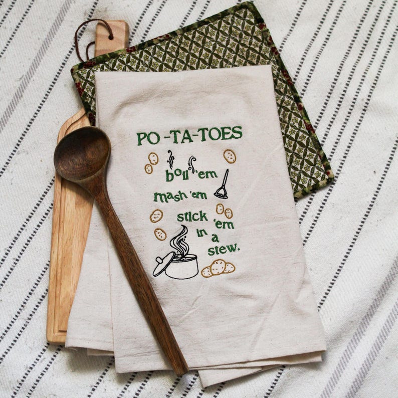 An unbleached off white linen tea towel embroidered with a quote from Lord of the Rings movie, the Two Towers. The Quote is Potatoes. Boil 'em, mash 'em, stick'em in a stew. Playful potatoes fall down to a  large whimsical pot, and a classic potatoes masher. The fonts represent the verbs, with steam coming off of the boil em phrase and the letters in mashed closely pushed together. Colors are a bold emerald green and a soft golden brown. Hang this towel in kitchen of any hobbit, elf, or wizard.