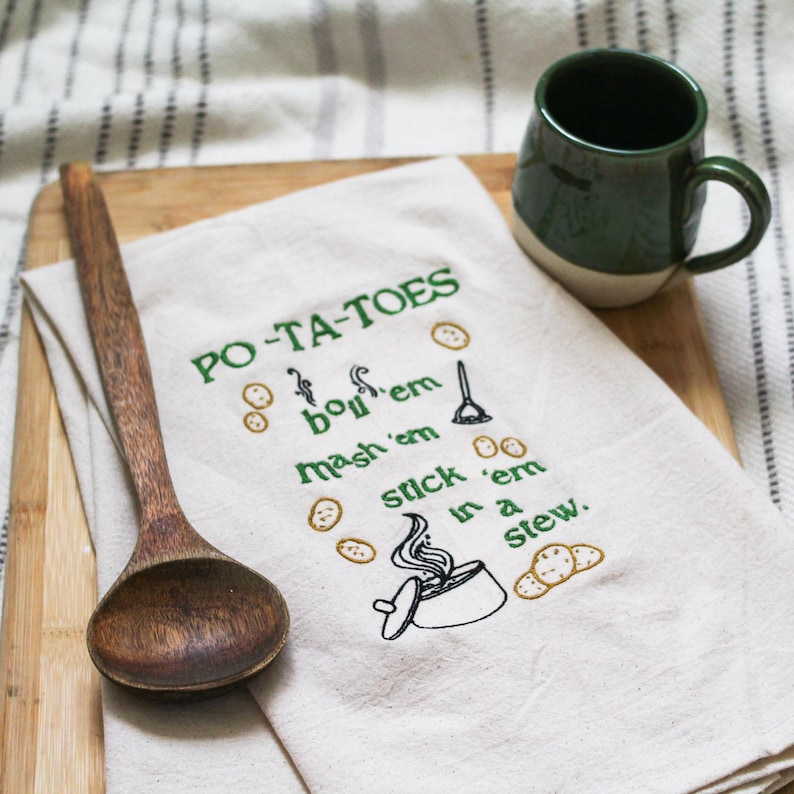 An unbleached off white linen tea towel embroidered with a quote from Lord of the Rings movie, the Two Towers. The Quote is Potatoes. Boil 'em, mash 'em, stick'em in a stew. Playful potatoes fall down to a  large whimsical pot, and a classic potatoes masher. The fonts represent the verbs, with steam coming off of the boil em phrase and the letters in mashed closely pushed together. Colors are a bold emerald green and a soft golden brown. Hang this towel in kitchen of any hobbit, elf, or wizard.